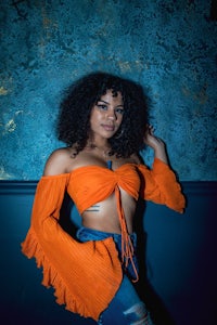 a black woman in an orange top posing against a blue wall